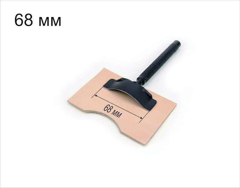 May include: A black leather working tool with a handle and a curved metal blade. The tool is placed on a piece of brown leather with a measurement of 68 mm marked on it.