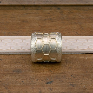 May include: A gold-colored metal ring with a hexagonal pattern. The ring is sitting on a light brown surface.