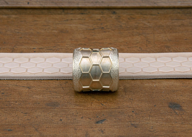 May include: A gold-colored metal ring with a hexagonal pattern. The ring is sitting on a light brown surface.