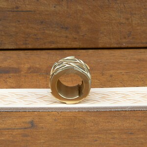 May include: A brass leather stamping roller with a geometric design. The roller is sitting on a piece of leather that has been stamped with the same design.