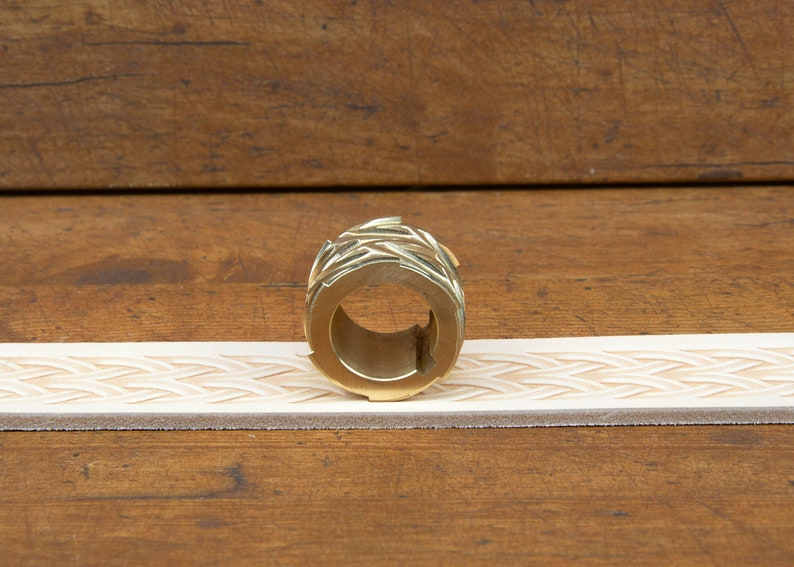 May include: A brass leather stamping roller with a geometric design. The roller is sitting on a piece of leather that has been stamped with the same design.