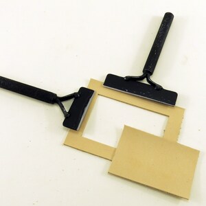 May include: Two black metal tools with handles are used to cut a rectangular piece of brown cardboard. The cardboard is cut to create a frame.
