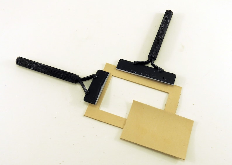 May include: Two black metal tools with handles are used to cut a rectangular piece of brown cardboard. The cardboard is cut to create a frame.