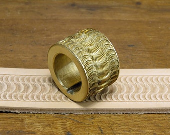 May include: A gold-colored leather stamping tool with a repeating pattern of overlapping semicircles. The tool is resting on a piece of leather that has been stamped with the same pattern.