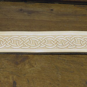 May include: A close-up of a light brown leather strip with a detailed Celtic knot design carved into the surface.