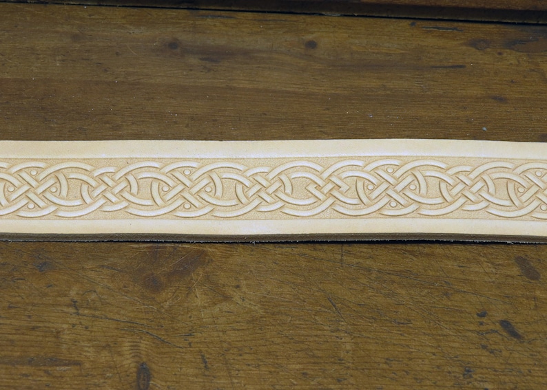 May include: A close-up of a light brown leather strip with a detailed Celtic knot design carved into the surface.