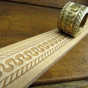May include: A close-up of a leather strip with a decorative pattern embossed into it. A brass tool with a similar pattern is shown rolling across the leather.