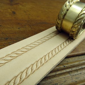 May include: A brass leather stamping tool with a rope design. The tool is being used to create a rope design on a piece of leather.