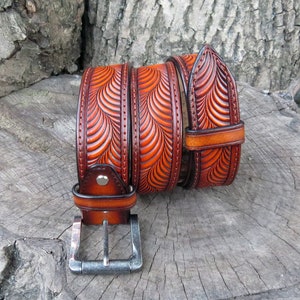 Pu&ograve; includere: Una cintura in pelle marrone con un intricato design intagliato di onde sovrapposte. La cintura ha una fibbia argentata ed &egrave; arrotolata su una superficie di legno.