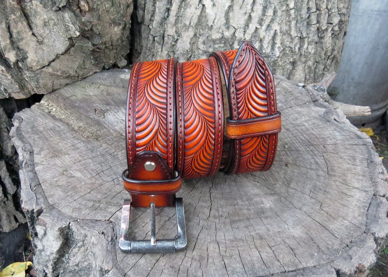 Pu&ograve; includere: Una cintura in pelle marrone con un intricato design intagliato di onde sovrapposte. La cintura ha una fibbia argentata ed &egrave; arrotolata su una superficie di legno.