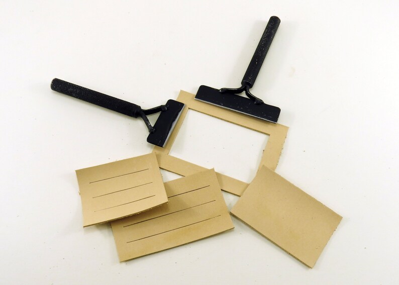 May include: Two black metal tools with handles and a flat blade, used for scraping or smoothing surfaces. The tools are lying on a white surface with four pieces of brown cardboard with lines on them.