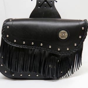 May include: A black leather saddlebag featuring a braided trim along the flap, silver-toned metal studs, and a long fringe detail. A circular silver-toned emblem with embossed text is centered on the front flap, which has a top mounting strap.