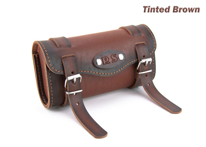May include: Tinted brown leather motorcycle saddlebag with two buckles and a metal plate with the initials "R.S." embossed on it.