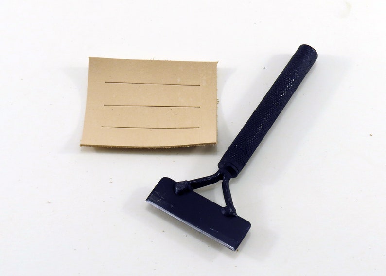 May include: A blue leather working tool with a handle and a blade. The tool is used to score leather. A piece of brown leather with lines scored into it is also shown.