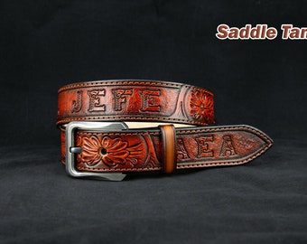 May include: A brown leather belt with the names "JEFE" and "AEA" embossed on it. The belt has a silver buckle and a floral design. The belt is labeled "Saddle Tan".