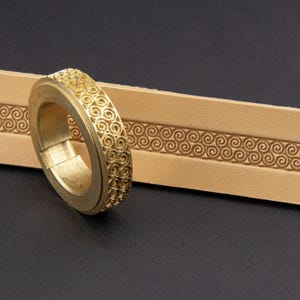 May include: A gold ring with a detailed spiral pattern around the band. Beside it is a tan leather strip, also featuring a spiral design. The background is a dark colour.