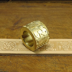 Könnte beinhalten: Ein goldfarbener Metallstempel mit einem detaillierten Design eines Lüfters und Zahnrädern. Der Stempel liegt auf einem Stück Leder, das mit dem gleichen Design geprägt wurde.