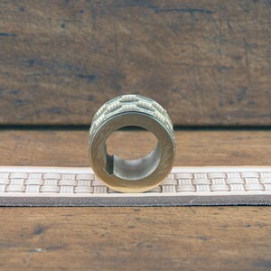 K&ouml;nnte beinhalten: Ein messingfarbener Metallring mit strukturierter, gemusterter Au&szlig;enseite. Der Ring hat eine zentrale &Ouml;ffnung und liegt auf einem hellen Lederg&uuml;rtel mit gewebtem Design. Der Hintergrund ist eine Holzoberfl&auml;che.