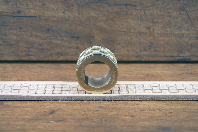 K&ouml;nnte beinhalten: Ein messingfarbener Metallring mit strukturierter, gemusterter Au&szlig;enseite. Der Ring hat eine zentrale &Ouml;ffnung und liegt auf einem hellen Lederg&uuml;rtel mit gewebtem Design. Der Hintergrund ist eine Holzoberfl&auml;che.