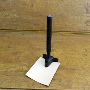 May include: A black leather punch tool with a handle and a square base is used to punch a hole in a piece of beige leather.