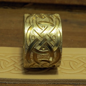 May include: A gold-colored metal stamp with a Celtic knot design. The stamp is used to create impressions in leather.