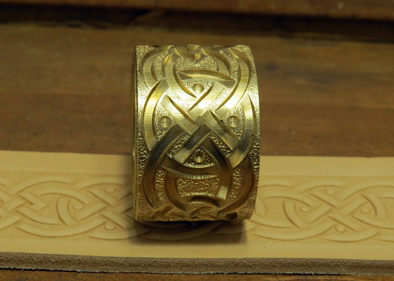 May include: A gold-colored metal stamp with a Celtic knot design. The stamp is used to create impressions in leather.