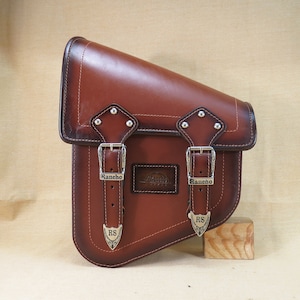 May include: Brown leather motorcycle saddlebag with two buckles and the word "Rancho" on each buckle. The bag has a brown leather strap with a silver buckle and the letters "RS" on it.