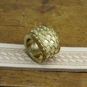 May include: A brass leather stamping tool with a woven pattern. The tool is on a piece of leather that has been stamped with the same pattern.