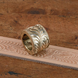 May include: A brass leather stamping tool with a floral design. The tool is lying on a piece of leather that has been stamped with the same design.