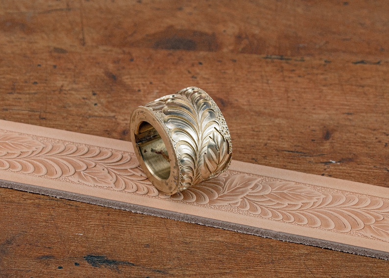 May include: A brass leather stamping tool with a floral design. The tool is lying on a piece of leather that has been stamped with the same design.