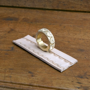 May include: A brass leather stamping tool with a decorative pattern of half circles. The tool is sitting on a piece of leather that has been stamped with the same pattern.