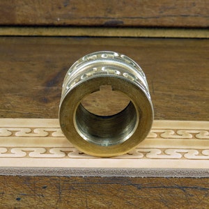 May include: A brass leather stamping roller with a decorative pattern. The roller is sitting on a piece of leather that has been stamped with the same pattern.