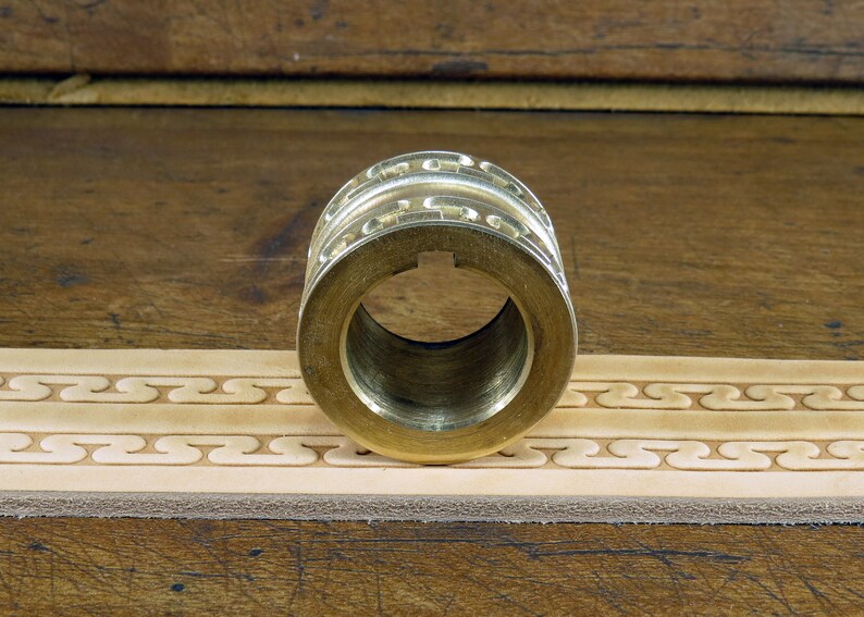 May include: A brass leather stamping roller with a decorative pattern. The roller is sitting on a piece of leather that has been stamped with the same pattern.