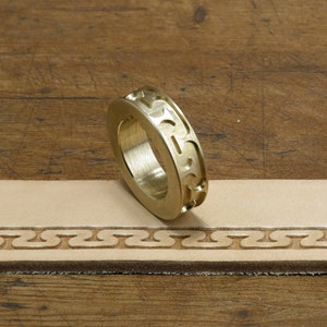 May include: A brass leather stamping tool with a decorative pattern. The tool is lying on a piece of leather that has been stamped with the same pattern.