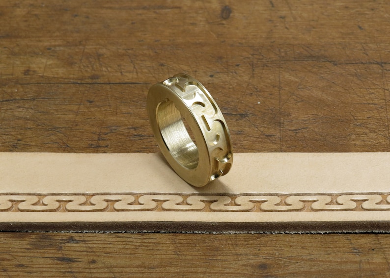 May include: A brass leather stamping tool with a decorative pattern. The tool is lying on a piece of leather that has been stamped with the same pattern.