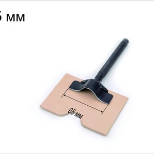May include: A black leather punch tool with a 65 mm diameter, shown on a piece of brown leather. The tool has a black handle and a metal base.