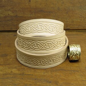 May include: Three leather belts with a Celtic knot design, stacked on top of each other. A gold metal belt buckle with a Celtic knot design is to the right of the belts.