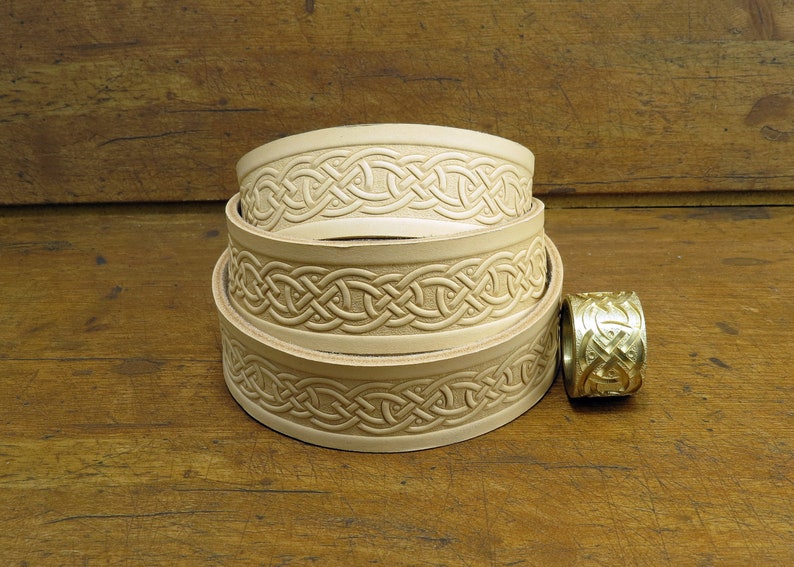 May include: Three leather belts with a Celtic knot design, stacked on top of each other. A gold metal belt buckle with a Celtic knot design is to the right of the belts.