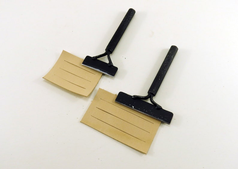 May include: Two black leather working tools with wooden handles, each resting on a piece of brown leather with parallel lines scored into it.