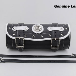 May include: Black leather motorcycle saddlebag with white trim and silver hardware. The bag has a buckle closure and a decorative gear design. The bag is labeled "Rancho" and comes with two black leather straps.