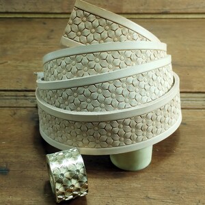 Peut inclure: Une pile de quatre lani&egrave;res de cuir blanc cass&eacute; avec un motif floral en relief sur la surface. Les lani&egrave;res sont enroul&eacute;es et pos&eacute;es sur une surface en bois. Un petit anneau en or avec un motif floral similaire se trouve au premier plan.