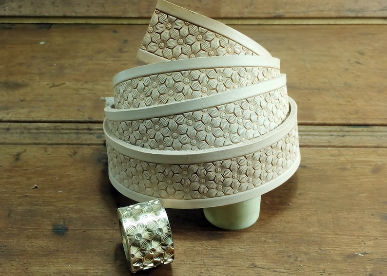 Peut inclure: Une pile de quatre lani&egrave;res de cuir blanc cass&eacute; avec un motif floral en relief sur la surface. Les lani&egrave;res sont enroul&eacute;es et pos&eacute;es sur une surface en bois. Un petit anneau en or avec un motif floral similaire se trouve au premier plan.