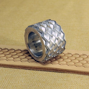 May include: A silver metal roller tool with a textured pattern of overlapping scales. The roller is resting on a piece of leather that has been embossed with the same pattern.