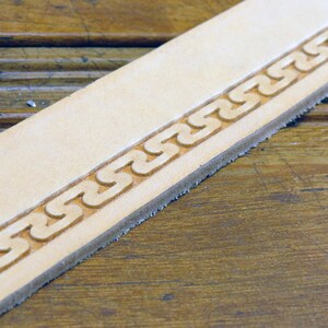 May include: Close-up of a light brown leather strip with a decorative border of a repeating chain pattern.
