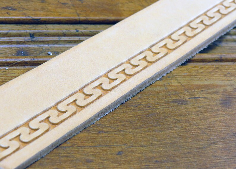 May include: Close-up of a light brown leather strip with a decorative border of a repeating chain pattern.