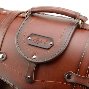Puede incluir: Una alforja de moto de cuero marr&oacute;n con asa superior y hebilla met&aacute;lica. La bolsa tiene un dise&ntilde;o cosido y una placa de metal con la palabra "Affolo" grabada.