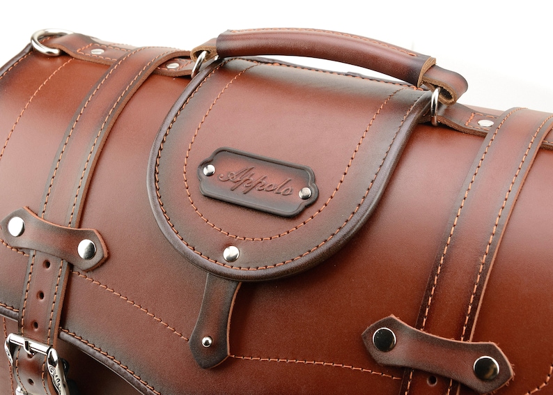 Puede incluir: Una alforja de moto de cuero marr&oacute;n con asa superior y hebilla met&aacute;lica. La bolsa tiene un dise&ntilde;o cosido y una placa de metal con la palabra "Affolo" grabada.