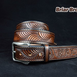 May include: A brown leather belt with a wave-like pattern and the word "NMAE" embossed on the tip. The belt has a silver buckle and is stitched with a contrasting thread.