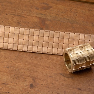 May include: A gold-colored ring with a woven pattern, next to a matching woven belt. The ring is cylindrical with a repeating square design. The belt is a light tan color and has a similar woven pattern. The items are on a wooden surface.