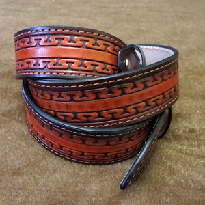 May include: A brown leather belt with a decorative pattern of interlocked lines. The belt is folded in half and the edges are stitched with a dark thread.